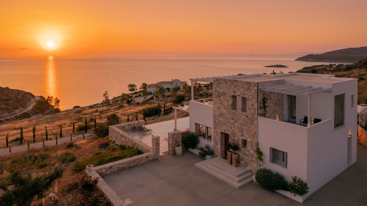 The two-story villa is showcased against a sunset over the Ionian Sea, with warm hues reflecting off the water. The exterior features a blend of modern and natural materials, surrounded by landscaped gardens. A spacious parking area is visible in the foreground.