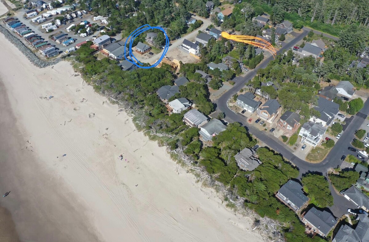 An aerial view captures Rockaway Cottage's location just back from the oceanfront, surrounded by lush greenery. The nearby beach and residential areas are visible, showcasing the cottage's proximity to the coastline and quiet neighborhood. The designated parking area is also in view.