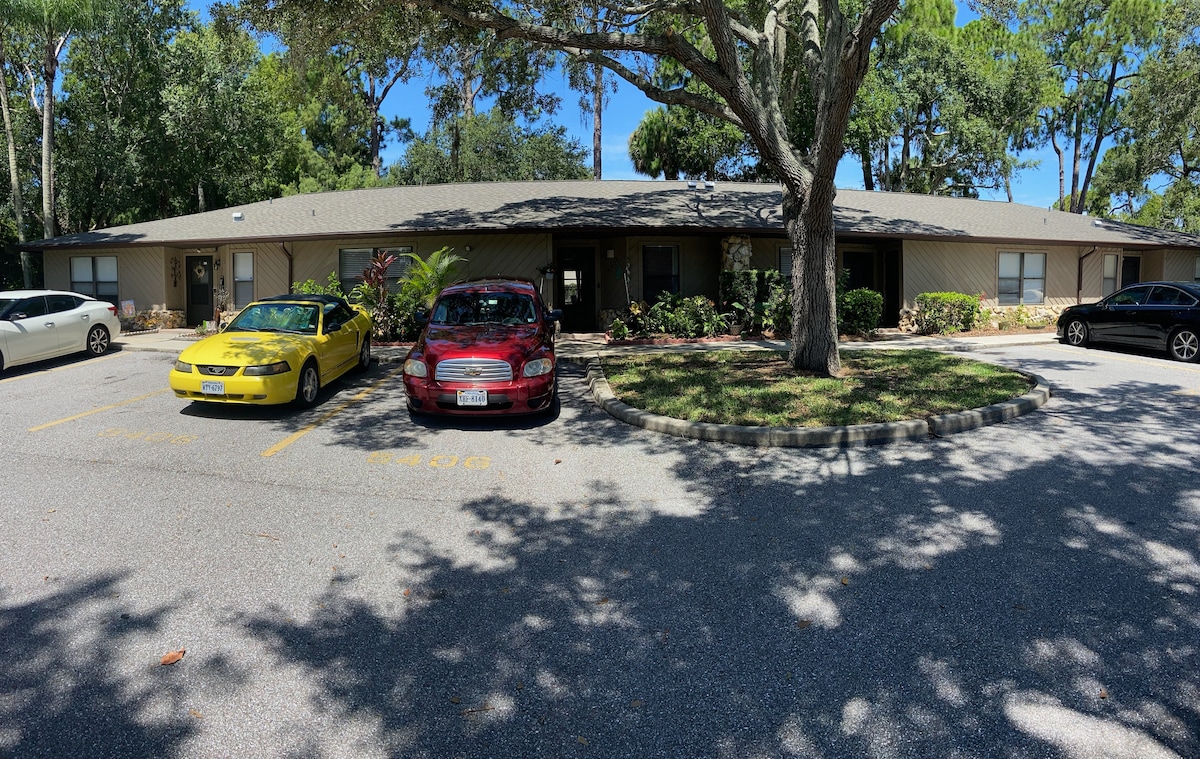 A view of the villa community reveals two parked vehicles—one yellow and one red—positioned in front of a single-story villa. Lush greenery and trees provide shade, while the building features a muted exterior with a modest front landscape.