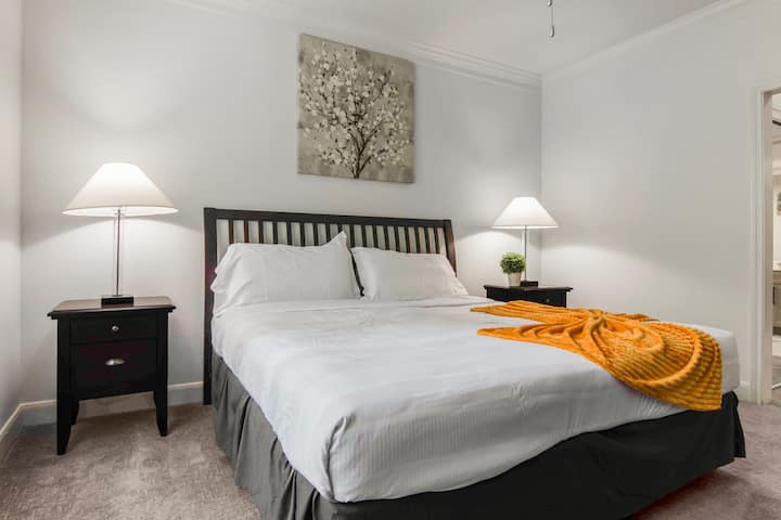 חדר שינה עם ראש מיטה מעץ, שתי שידות לילה עם מנורות, רצפת שטיחים ופלטת צבעים נייטרלית עם נגיעות צבע צהובות.