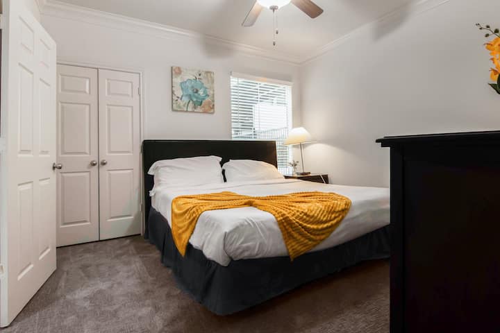 חדר שינה עם ראש מיטה כהה, מיטה לבנה עם שמיכה צהובה, רצפת שטיחים, מאוורר תקרה ודלתות ארון כפולות.