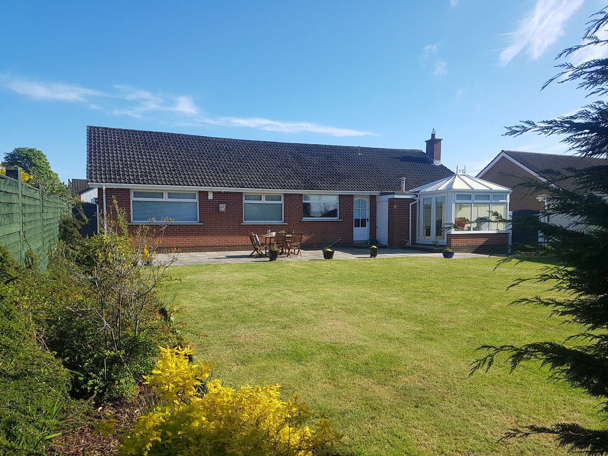 A spacious bungalow is showcased, featuring a light exterior and set against a clear blue sky. A large green garden surrounds the property, complemented by a patio area with seating and a glass conservatory, enhancing outdoor relaxation.