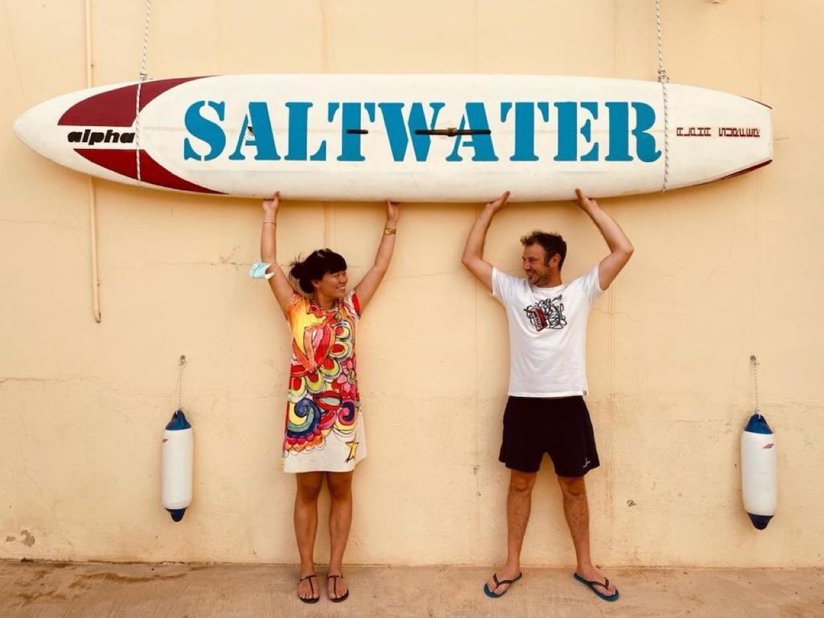 A surfboard labeled 'SALTWATER' is displayed prominently against a wall. Two individuals pose underneath the board, showcasing a colorful beach outfit and casual attire. The setting conveys a relaxed atmosphere, with elements suggestive of a seaside theme.