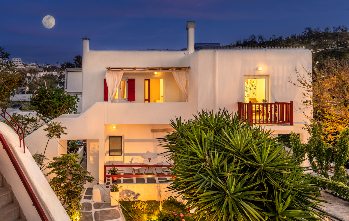 A charming two-story structure is illuminated under the moonlight, surrounded by lush greenery. Balconies provide an outdoor view, while large windows invite natural light into the interior. The garden is well-maintained, enhancing the tranquil ambiance of the setting.