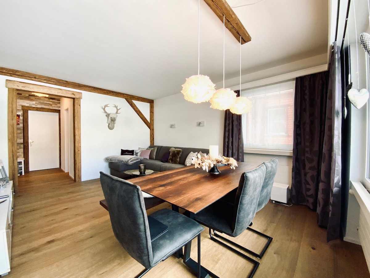 A spacious living area is presented with a wooden dining table surrounded by dark chairs. Soft pendant lights hang above the table, while a cozy sofa with decorative cushions is visible along one wall. Natural light filters through large windows covered with sheer curtains.