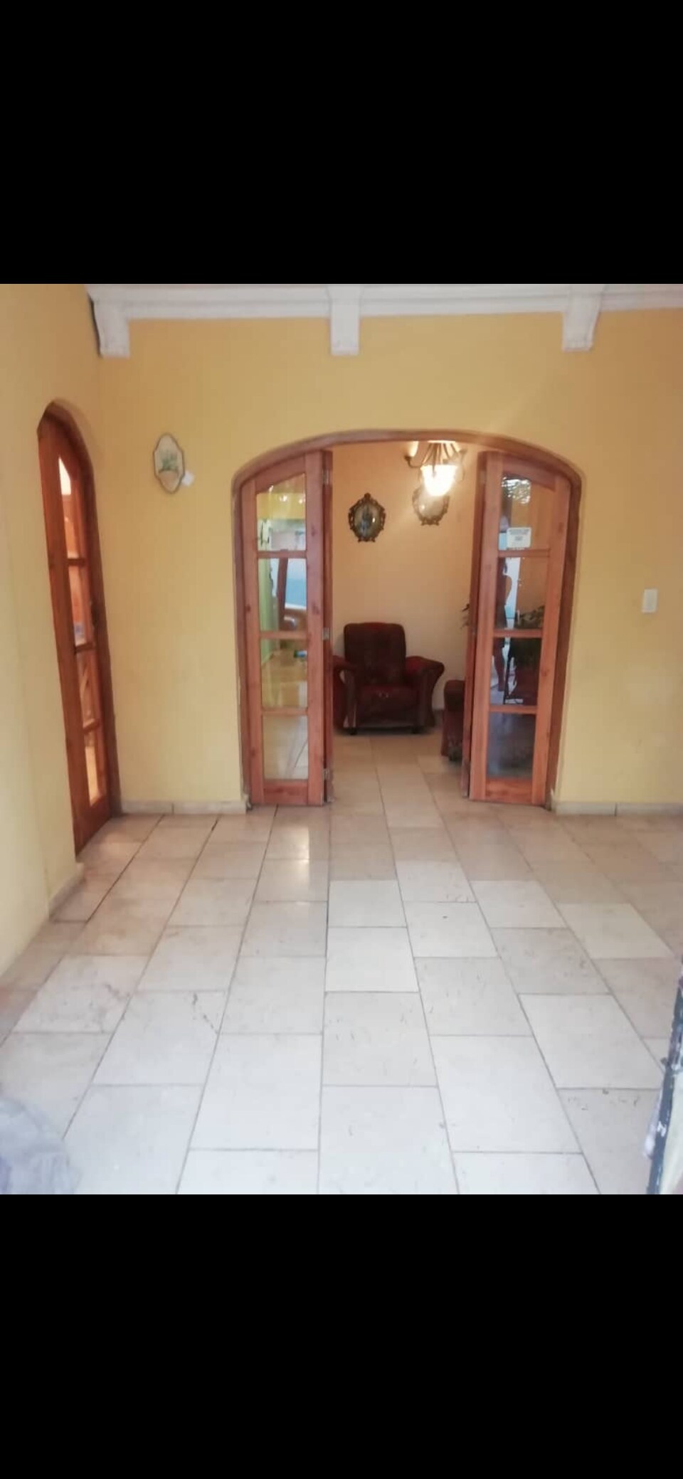 An entrance area showcases a tiled floor and is framed by two wooden archways. Light wooden doors lead to a cozy seating area featuring a pair of armchairs. The walls are painted a soft yellow, enhancing the bright ambiance.