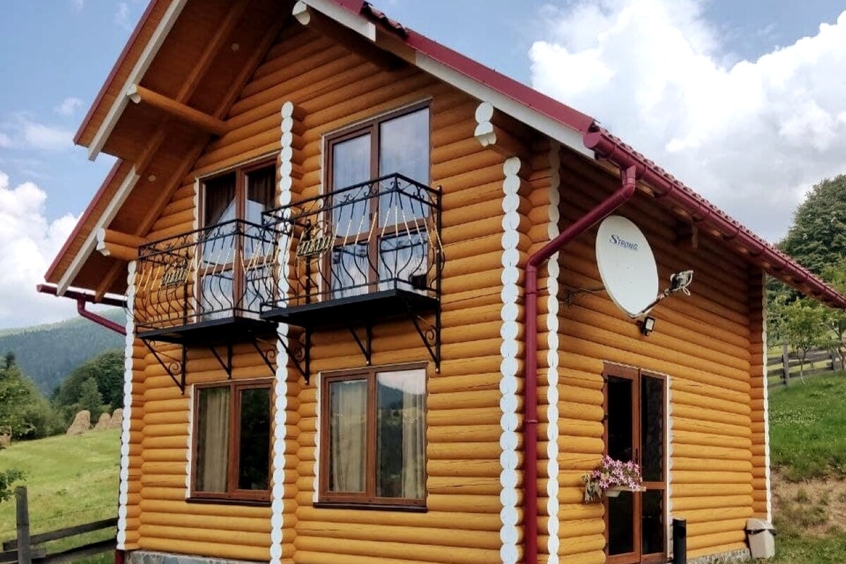A wooden house with a charming exterior features two balconies adorned with decorative railings. Large windows allow natural light to fill the rooms, while a satellite dish is mounted on the side. The surrounding landscape showcases grassy slopes and gentle hills.