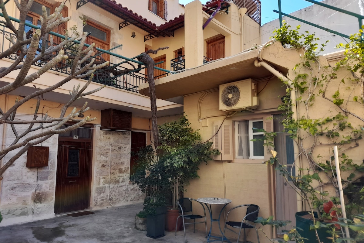 A shared outdoor courtyard is visible, featuring a small table and chairs surrounded by greenery. A wall-mounted air conditioning unit is present, and a low roof extends over part of the area. The entrance to the building is partially seen, showcasing traditional stone architecture.