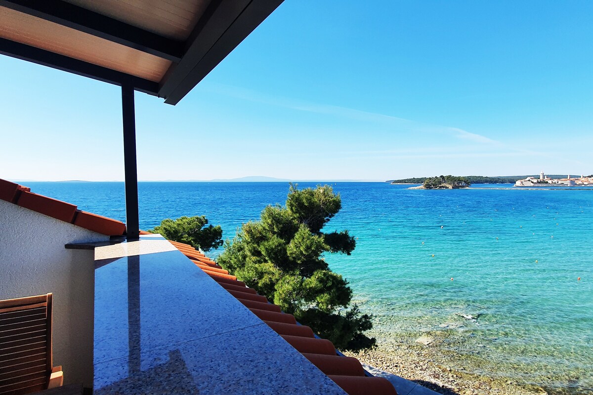 An inviting view of the clear blue sea is captured from a balcony. The coastline is framed by green trees, with the tranquil water stretching toward distant islands under a clear sky.
