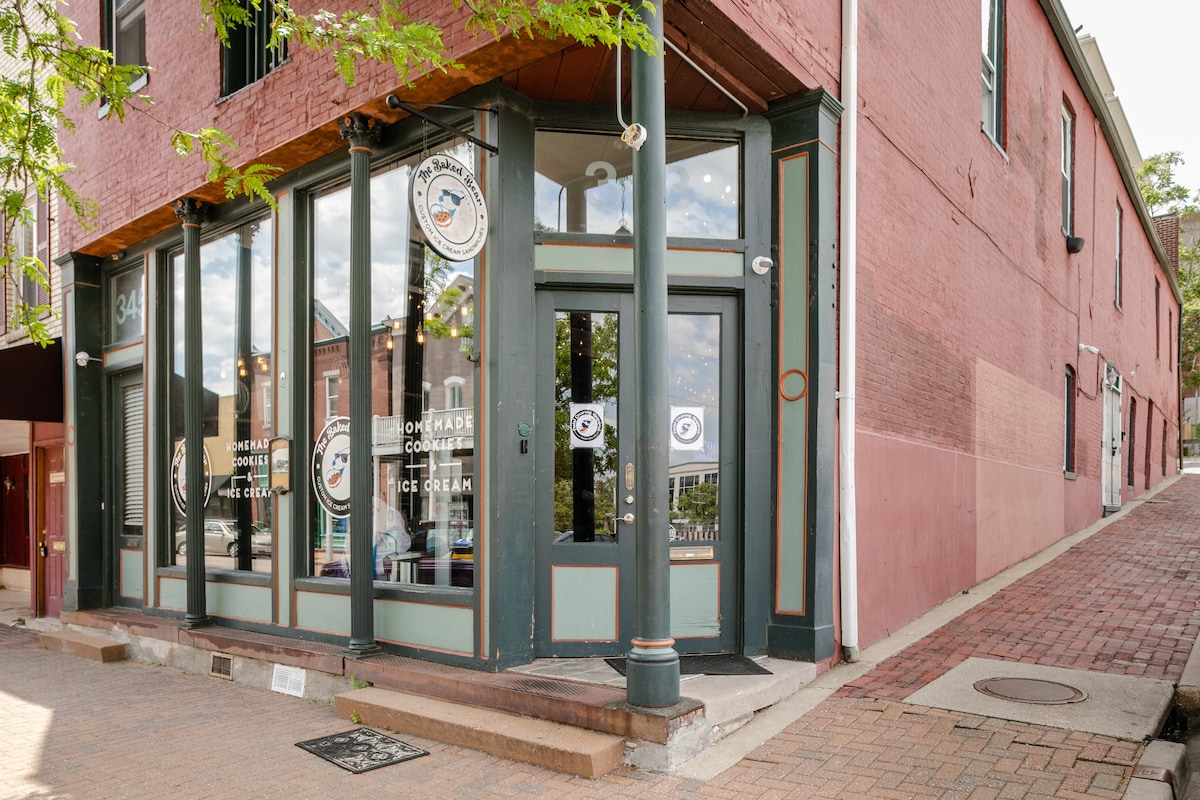 The exterior of a historic building is visible, featuring large glass doors framed by green trim. The facade is painted in warm earthy tones, complemented by large windows displaying the interior. The street slopes gently, and a sign indicating the presence of a nearby ice cream shop can be seen.