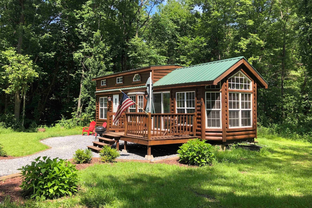 The cabin features a wooden exterior with a green metal roof, surrounded by trees. A welcoming porch with railings extends from the front, complete with red chairs and an American flag. The landscaped yard includes well-maintained greenery and gravel pathways.