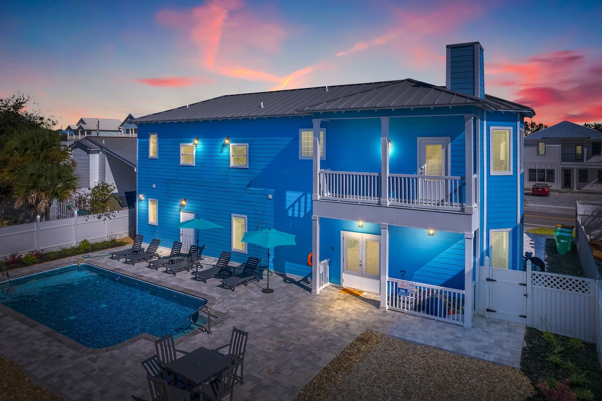 The exterior of the property showcases a vibrant blue facade, complemented by a spacious backyard featuring a swimming pool. Lounge chairs surround the pool, and a seating area with a table is positioned nearby. The two-story structure is illuminated by soft lighting at dusk.
