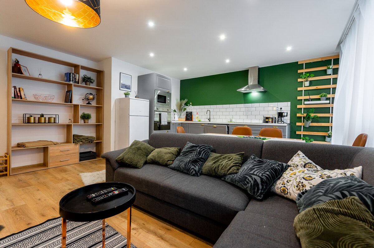 A modern living area is presented with a large gray sectional sofa adorned with various decorative cushions. Open shelving displays books and plants, while a fully equipped kitchen can be seen in the background. Natural light enhances the green accent wall.