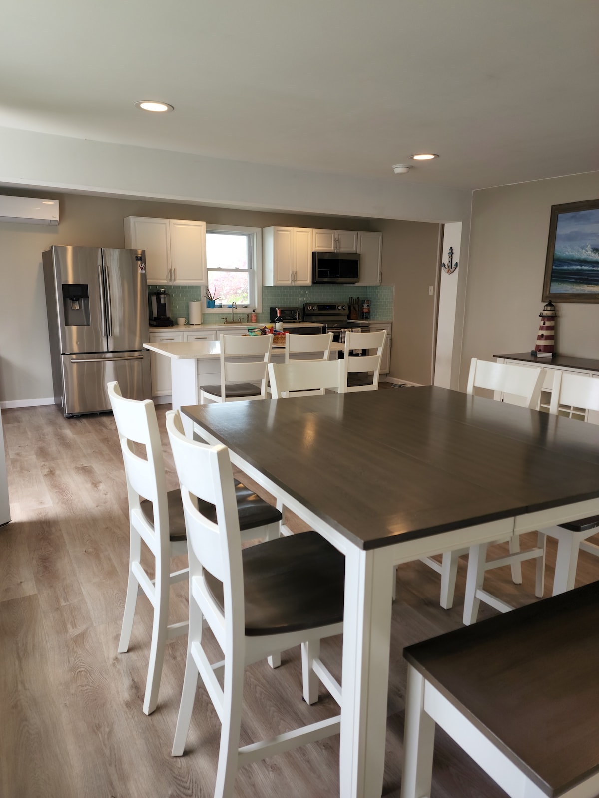 An open concept kitchen and dining area is highlighted by a large table with eight white chairs. Modern appliances, including a stainless steel refrigerator and oven, are visible in the background. Natural light illuminates the space through a nearby window.