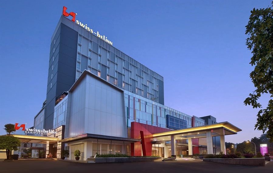 The hotel exterior features a modern architectural design with a combination of glass and metal elements. The entrance is highlighted by a spacious overhang and illuminated signage, while the surrounding landscape includes well-maintained greenery. Evening lighting enhances the building's inviting atmosphere.
