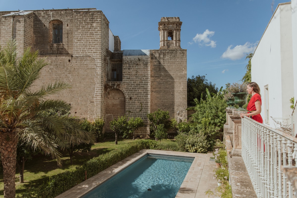 A serene outdoor space features an inviting pool set against the backdrop of ancient stone walls and a historic tower. Lush greenery surrounds the area, enhancing the peaceful ambiance of the garden. A pathway leads to the poolside, providing access to the tranquil setting.