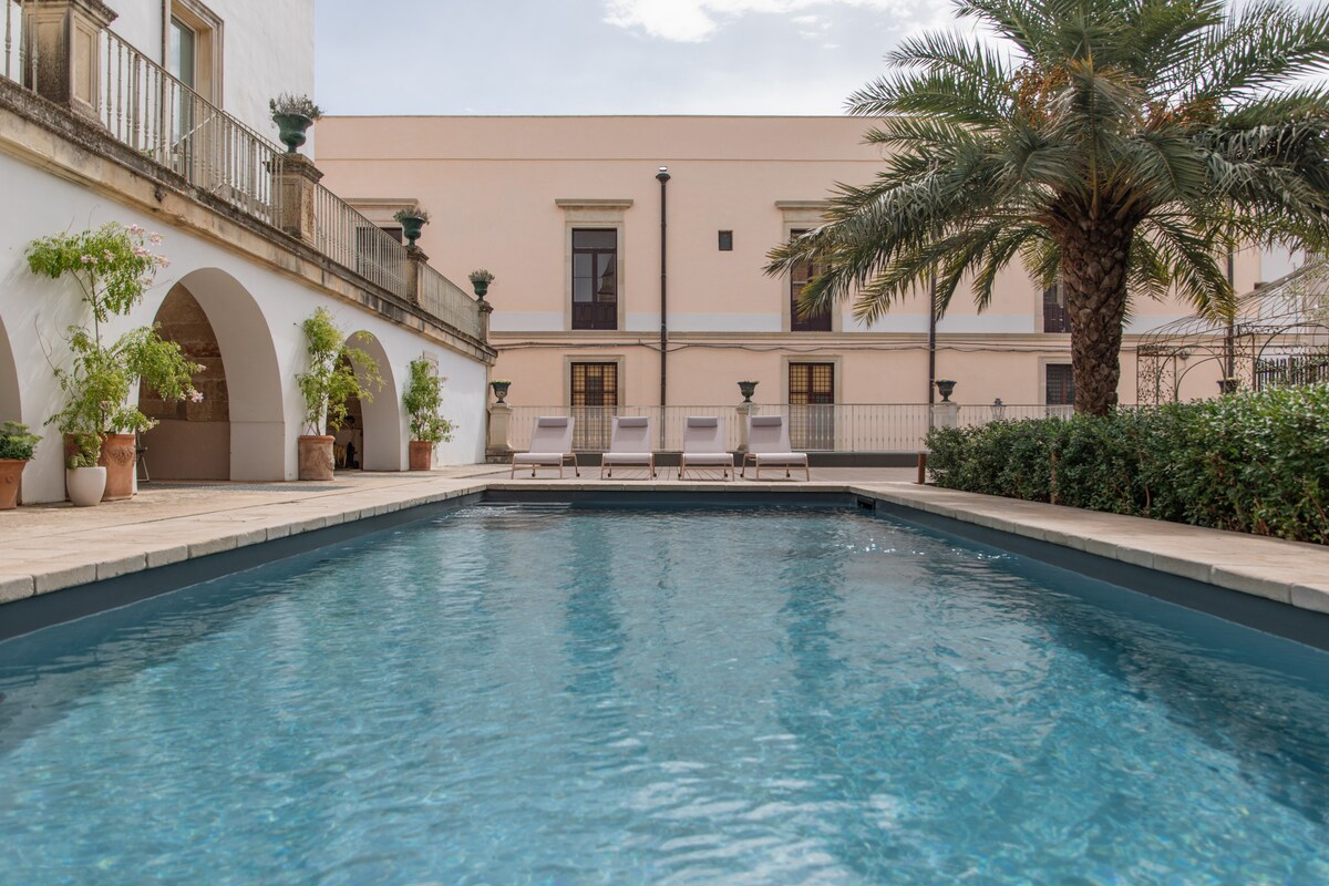 An outdoor swimming pool is surrounded by a stone patio, with palm trees and shrubs providing greenery. Sun loungers are positioned along one side of the pool, with an elegant building visible in the background, featuring arches and balcony areas.
