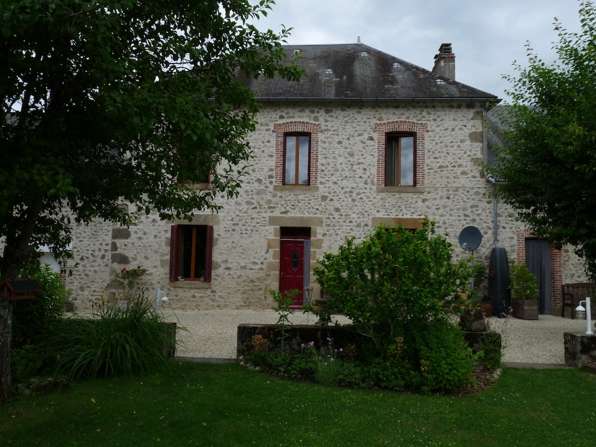 The front view of a charming stone house features a gravel path leading to an inviting entrance. Large windows with red trim are visible, framed by greenery. A garden area showcases lush grass and shrubs, with a few flowering plants along the path.