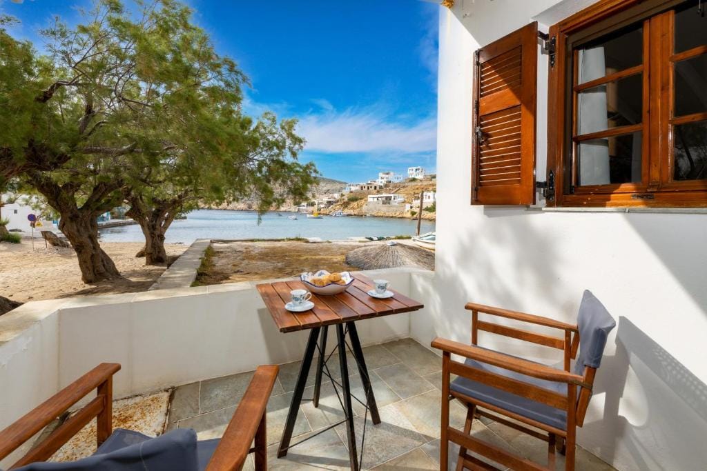 An outdoor seating area features a small wooden table surrounded by two chairs, offering views of the nearby water. Lush greenery is visible to one side, and a partially open window adds to the inviting ambiance of the space.