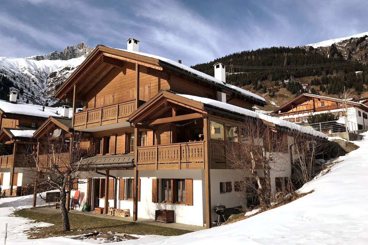 A wooden chalet-style building is set against a mountainous backdrop, with snow-covered ground and rooftops. Several balconies, framed by large windows, offer outdoor seating. Surrounding trees and a small yard complement the scenic environment.