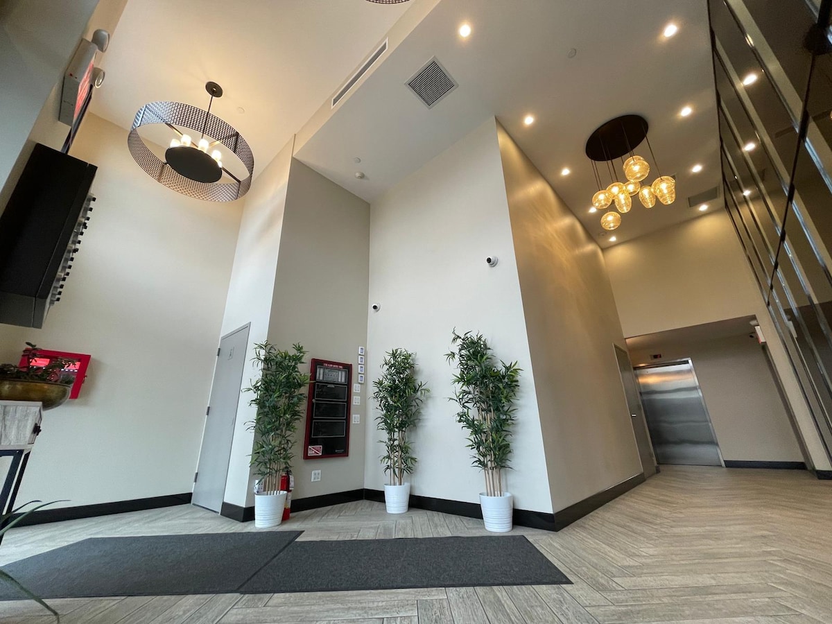 An elevator lobby features high ceilings illuminated by modern light fixtures. The floor is covered with a stylish tile design, and two potted plants add natural elements. A wall-mounted directory and an elevator bank are visible, providing easy navigation for guests.