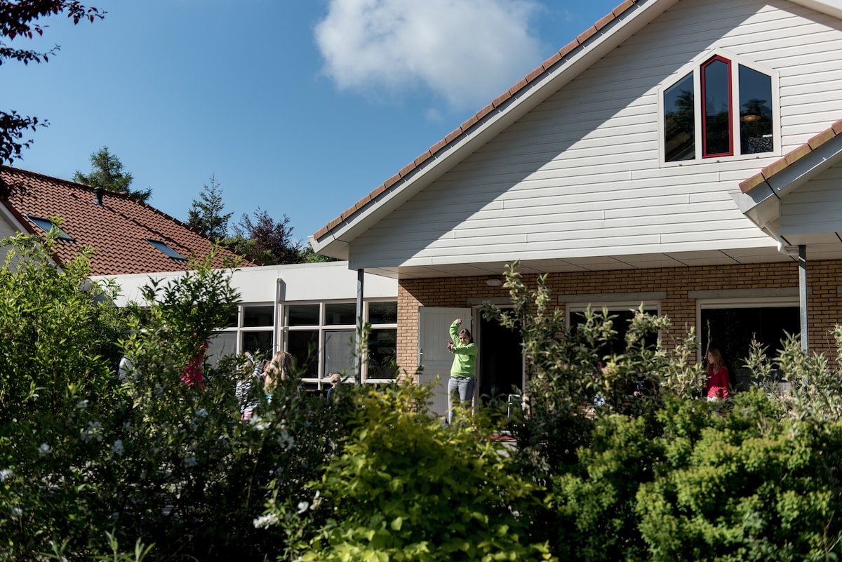 A well-maintained building with a sloped roof is visible, surrounded by lush greenery and colorful flowering plants. Large windows reflect sunlight, enhancing the inviting appearance of the space. People can be seen enjoying the outdoors, contributing to a lively atmosphere.