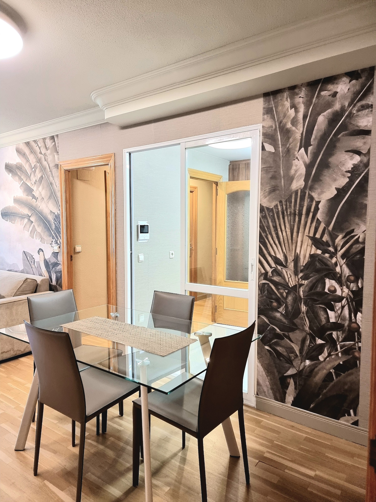 A modern dining area is featured, showcasing a glass-top table surrounded by four sleek chairs. The walls are adorned with artistic botanical prints, and a doorway can be seen in the background, leading to another room.