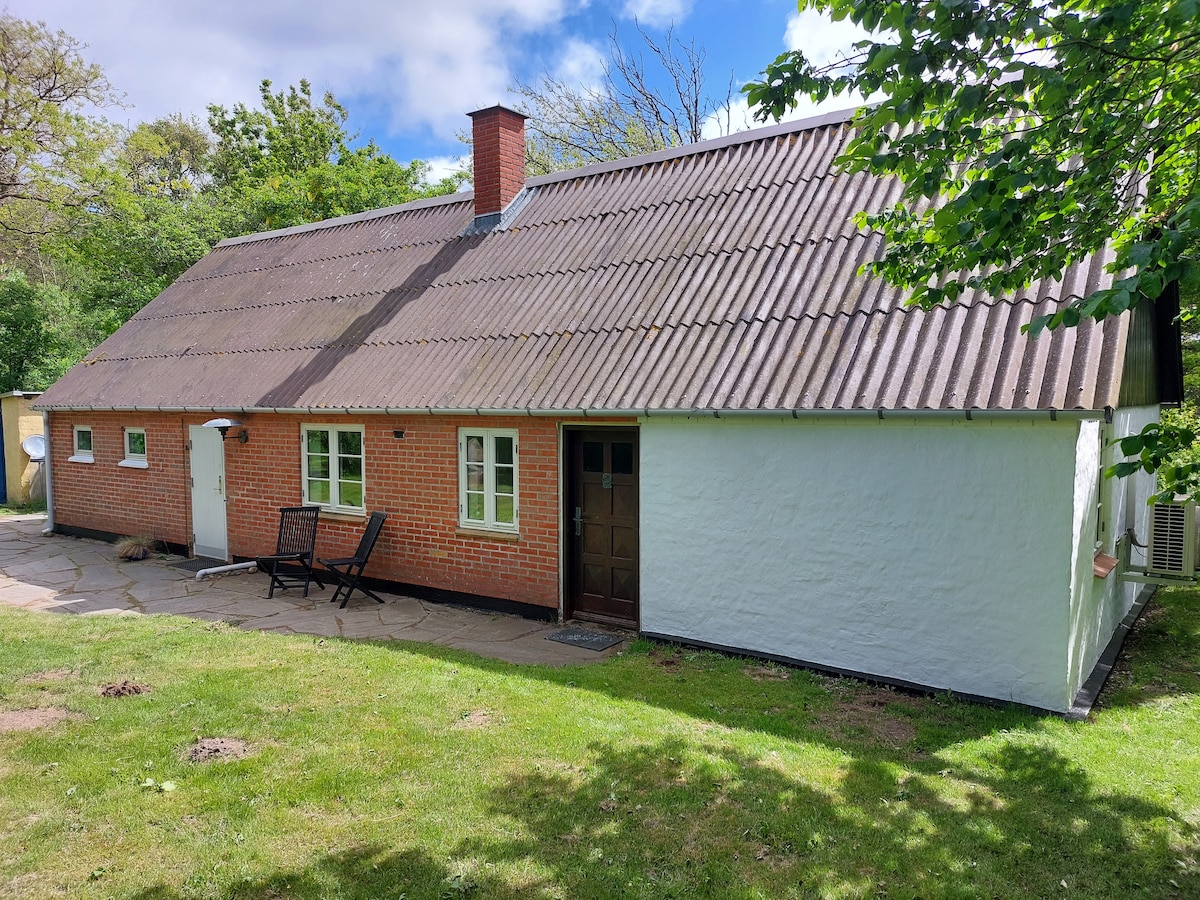 The exterior of a charming country house is shown, featuring a sloped roof and a mix of brick and white walls. A small patio area with two chairs is visible, surrounded by greenery and landscaped lawn. Trees are present in the background, contributing to a tranquil setting.