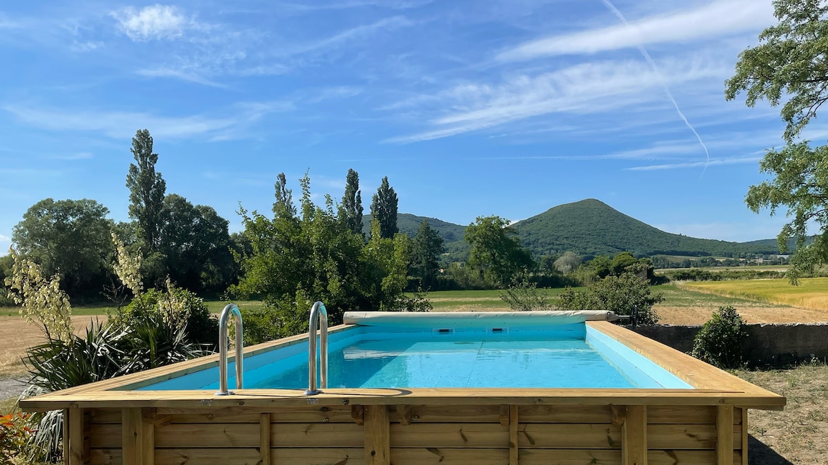 A wooden pool deck surrounds a secure above-ground pool, set against a backdrop of rolling green hills and clear blue skies. Lush trees frame the scene, hinting at the tranquil outdoor space available for relaxation.