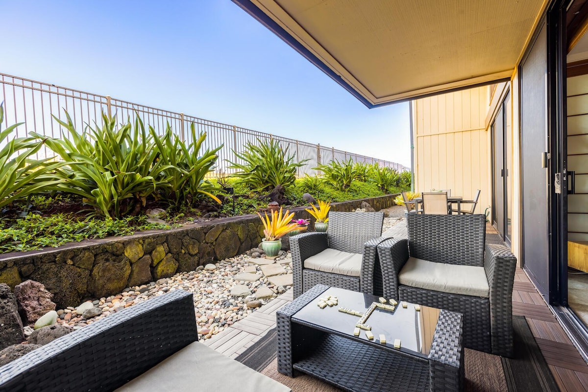 An outdoor seating area is furnished with modern wicker chairs and a coffee table, situated under a shaded awning. Lush greenery surrounds the space, with colorful plants and smooth stones creating a serene garden ambiance.