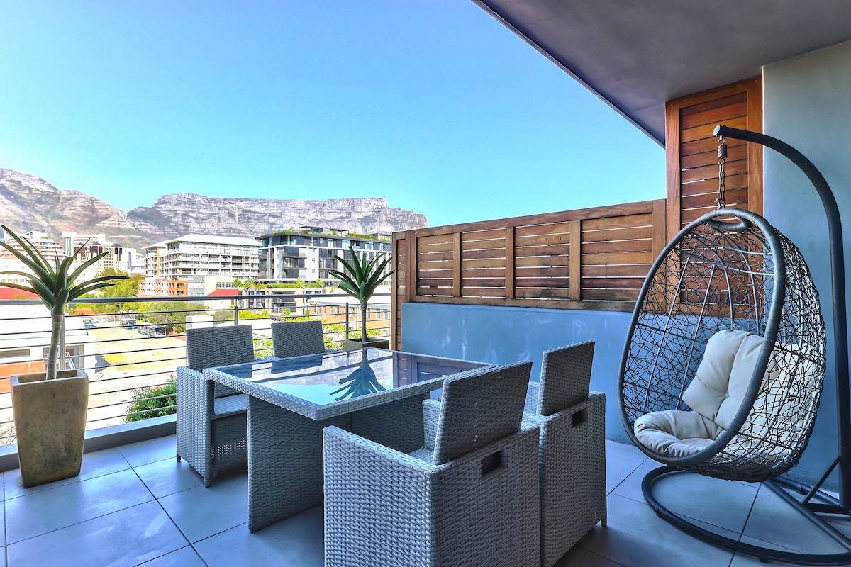 A spacious balcony is presented, featuring a glass dining table surrounded by four woven chairs. A hanging chair adds an inviting touch, while potted palms enhance the outdoor space. Majestic Table Mountain provides an impressive backdrop under a clear blue sky.