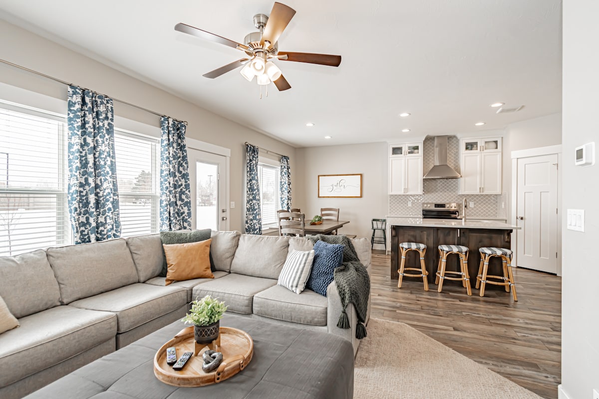 A spacious living area features a large sectional sofa adorned with decorative pillows. Light streams through large windows, complemented by patterned curtains. A round coffee table sits on a wooden floor, and a dining area with sleek stools is visible in the background, creating a harmonious gathering space.