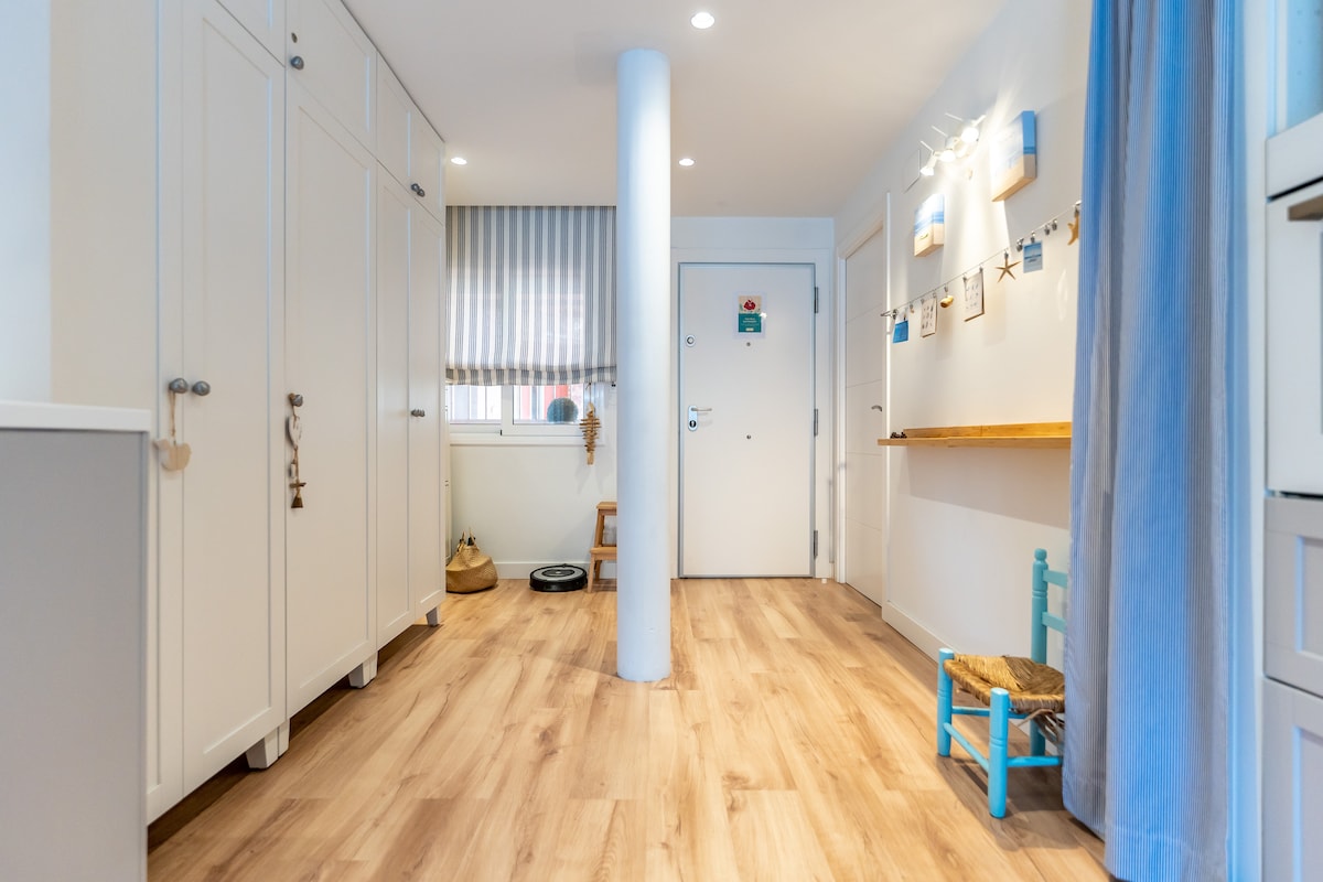 A spacious hallway welcomes guests with light wood flooring and soft, neutral tones. Storage cabinets line one side, while a small blue chair and a decorative shelf with nautical-themed items are positioned on the other. Natural light filters through a striped curtain adorning the window.