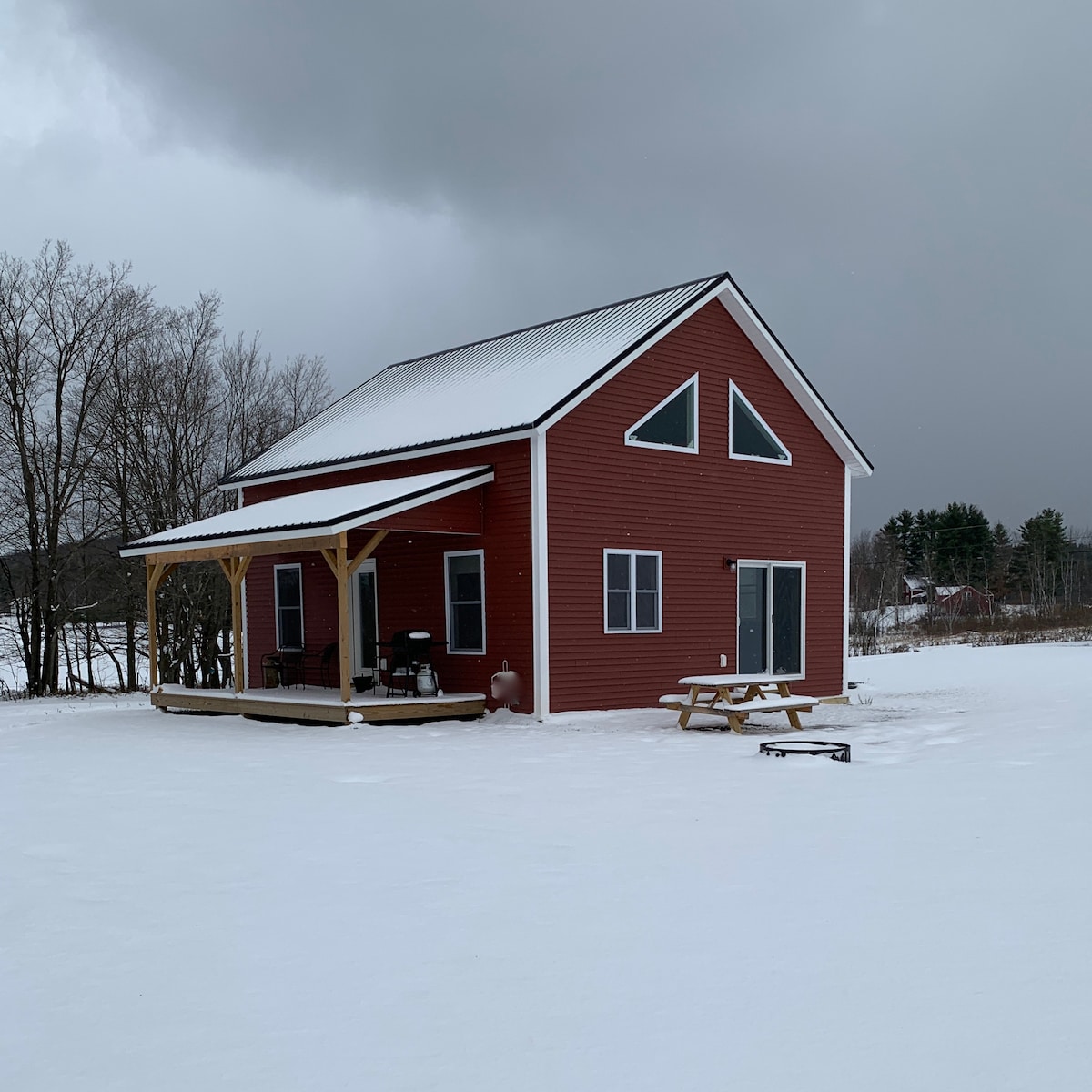 A red cottage is shown, featuring a porch supported by wooden posts. Snow covers the ground surrounding the structure, and large windows allow light to enter. A picnic table is positioned on the front deck, with a grill visible nearby, set against a backdrop of trees.