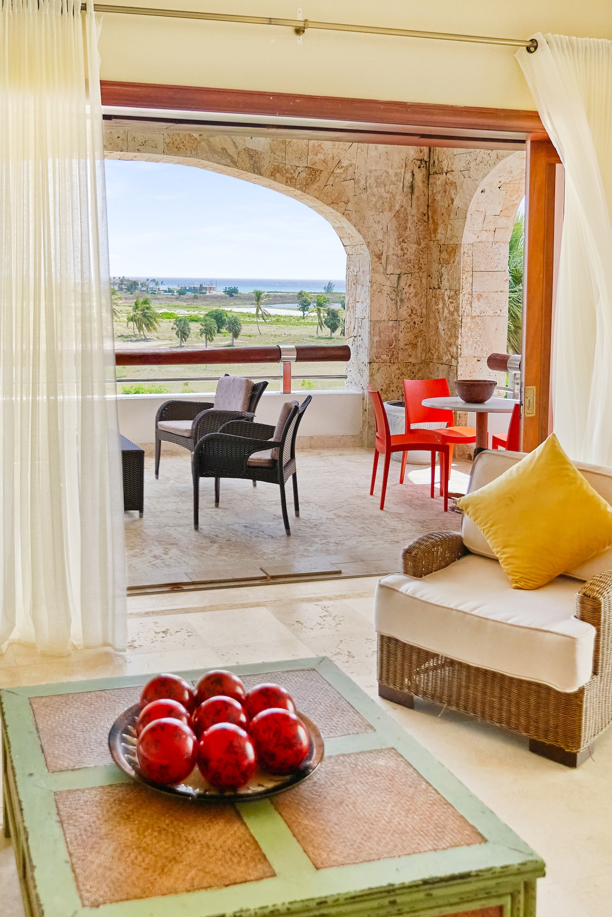 A spacious living area features a comfortable seating arrangement with a plush sofa and vibrant accents. An outdoor balcony is visible, furnished with a red table and chairs, offering expansive views of the landscape and sea. Light filters through sheer curtains, creating a bright ambiance.