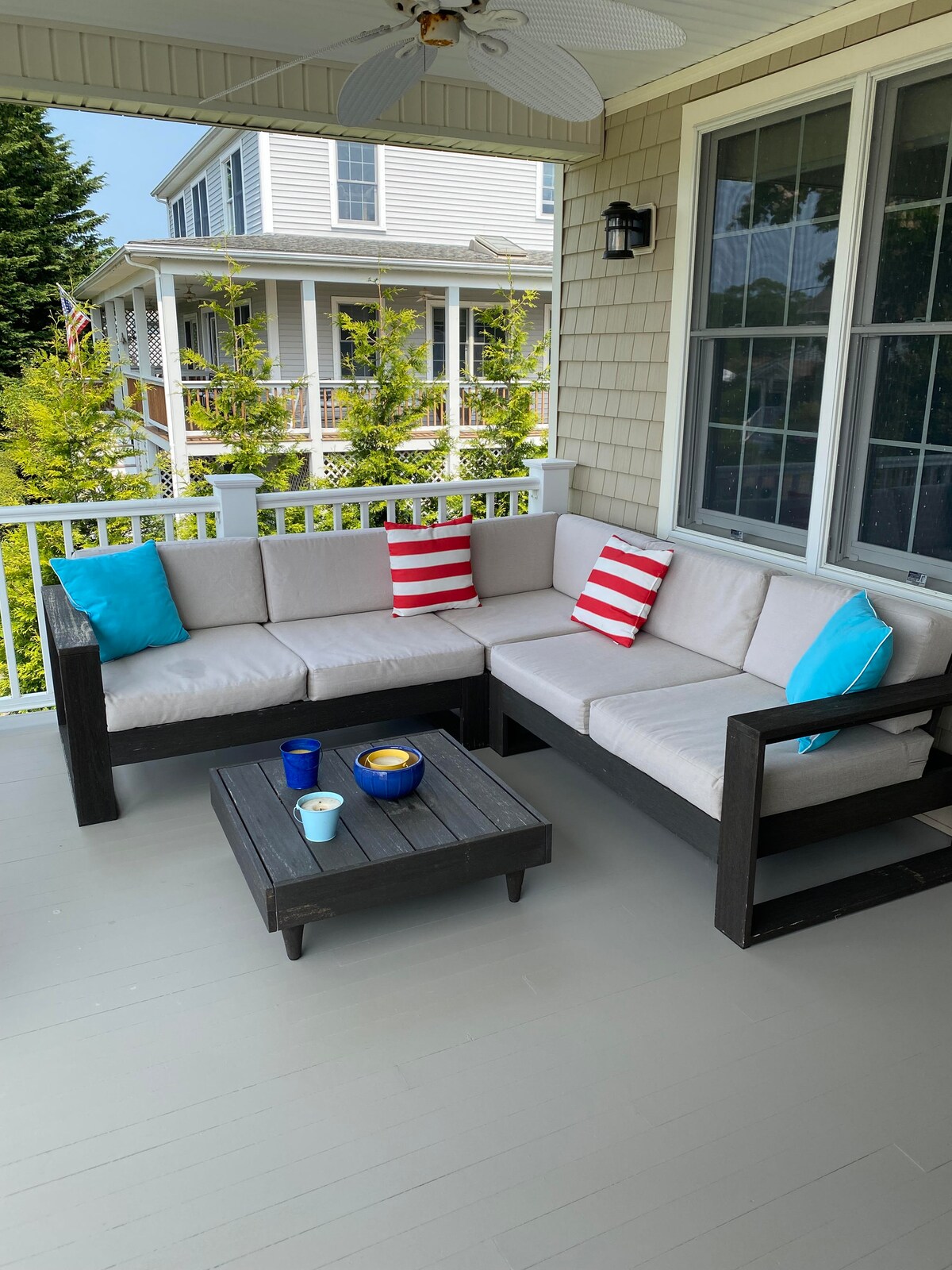 A sectional seating area is arranged with light-colored cushions and decorative pillows in blue and red stripes. A dark wooden coffee table holds snacks and drinks, complementing the relaxed outdoor setting. Sunlight streams through the nearby windows, enhancing the inviting space.