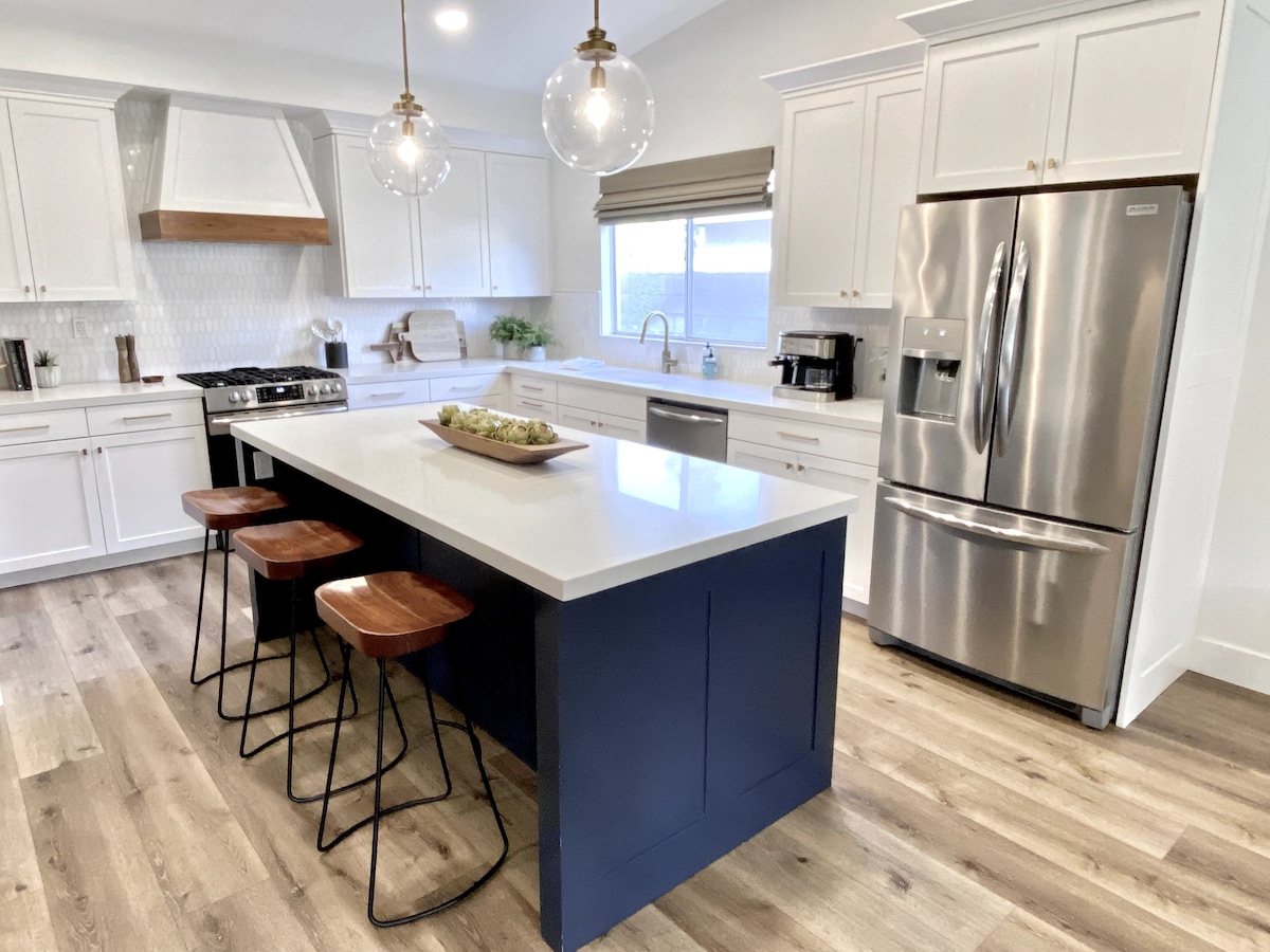 A modern kitchen is highlighted with stainless steel appliances and white cabinetry. A large island featuring a blue base provides seating for three on wooden stools. Natural light floods in from the window above the sink, accentuating the clean lines and open space.
