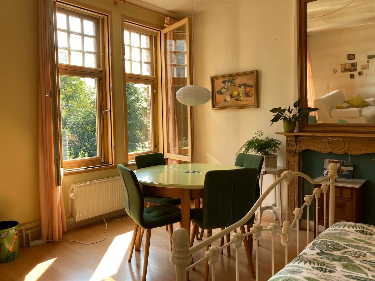 A bright dining area features a round yellow table surrounded by green upholstered chairs, illuminated by natural light from large windows. A simple pendant light hangs above the table, and a decorative green plant is positioned nearby, adding a touch of greenery to the space.
