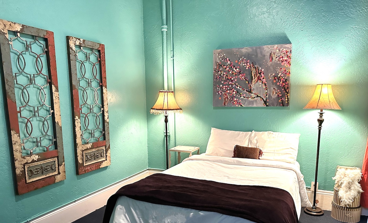 A cozy room features a spacious bed adorned with a dark blanket. Two unique lamps illuminate the space, highlighting teal walls. Decorative panels flank the bed, and a floral artwork adds a touch of color above. Soft light enhances the welcoming atmosphere.