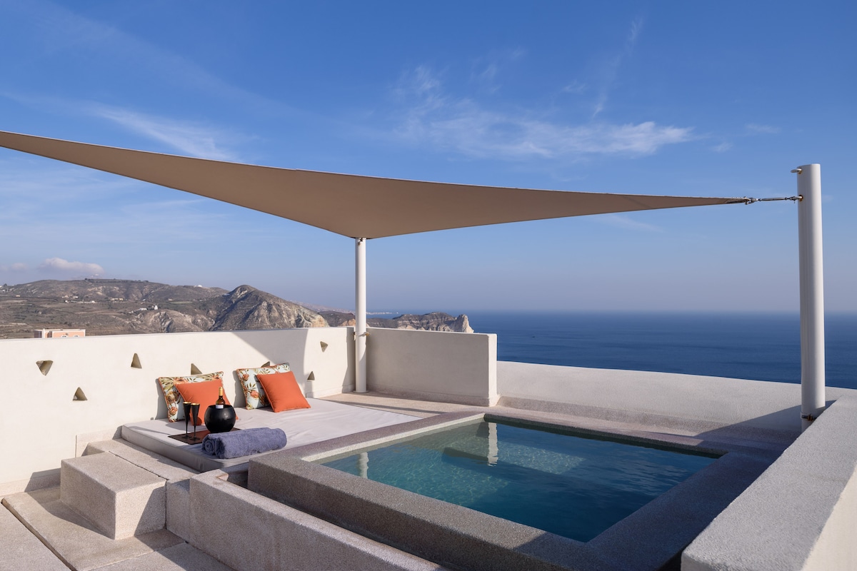 A large private veranda features a shaded seating area with decorative pillows and a coffee table, overlooking the Aegean Sea. A small plunge pool is situated nearby, reflecting the clear blue sky and the serene horizon, enhancing the space for relaxation.