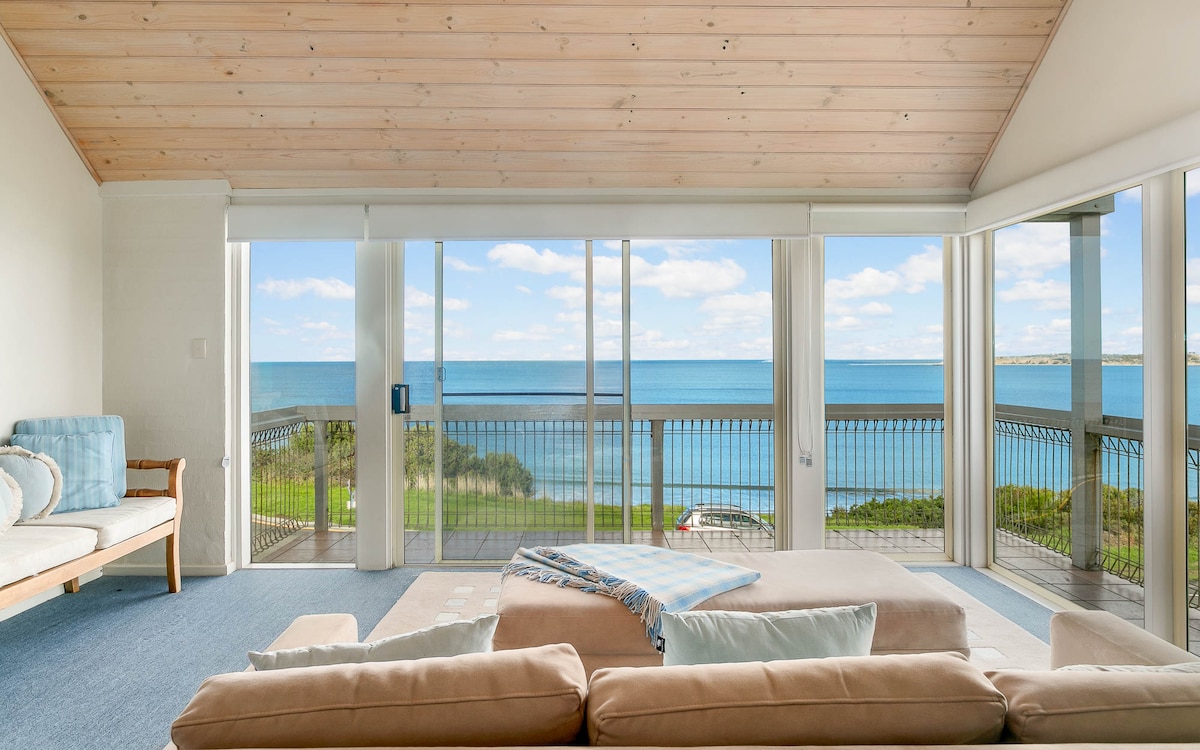 A spacious living area is highlighted by large glass doors that open to panoramic ocean views. Soft neutral furnishings are arranged around a central area, offering a comfortable place to relax. Natural light fills the space, creating a calm atmosphere ideal for unwinding.