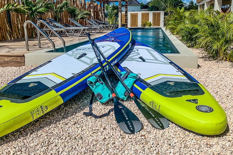 Two stand-up paddle boards lie partially on a pebble surface, accompanied by paddles and a blue accessory holder. In the background, a magnesium lap pool is visible along with lounge chairs, surrounded by tropical greenery.
