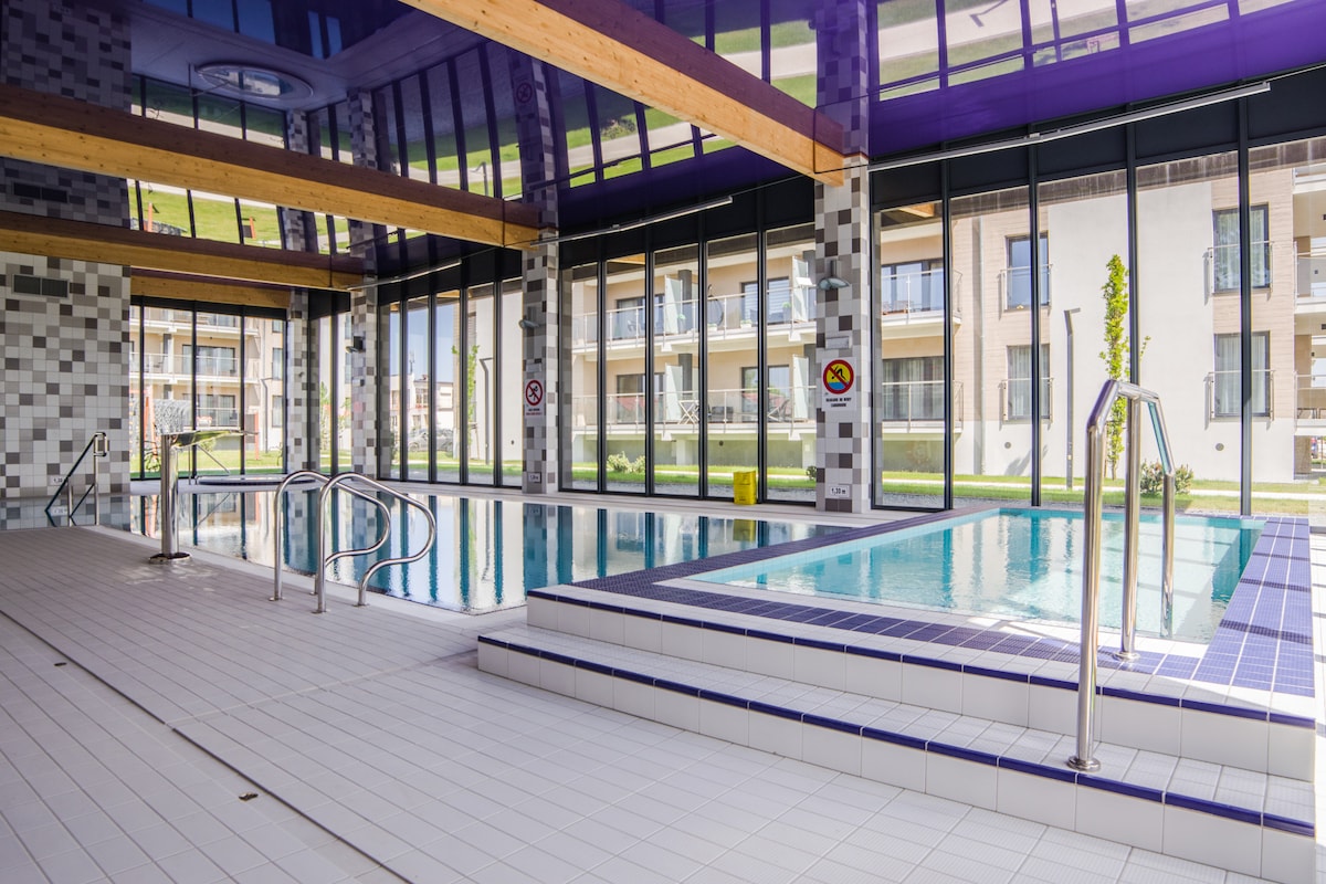 An indoor pool area is featured, surrounded by large glass windows that offer ample natural light. The space includes both a swimming pool and a jacuzzi, with a tiled terrace leading to the water. Wooden beams add structural interest to the high ceiling.