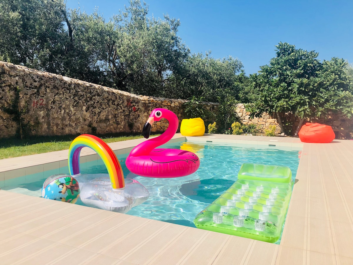 The private pool area features colorful inflatables, including a pink flamingo and a rainbow ring, surrounded by a spacious sunbathing area. Bright sunshine reflects off the water, while vibrant beanbags provide additional seating options, blending relaxation with outdoor fun in a tranquil garden setting.