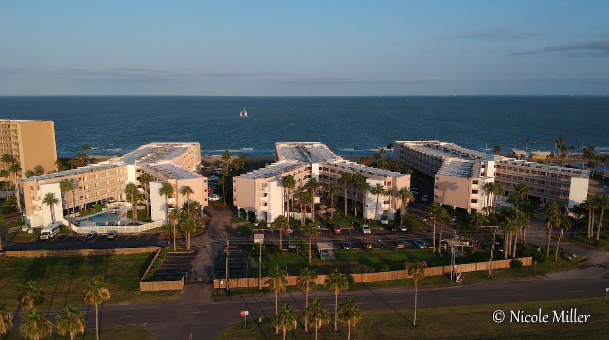 An aerial view captures the gated Villa del Sol complex, featuring multiple connected buildings surrounded by palm trees. The beach and ocean are visible beyond the property, with a sailboat on the horizon. A swimming pool is seen within the complex, enhancing the aquatic ambiance.
