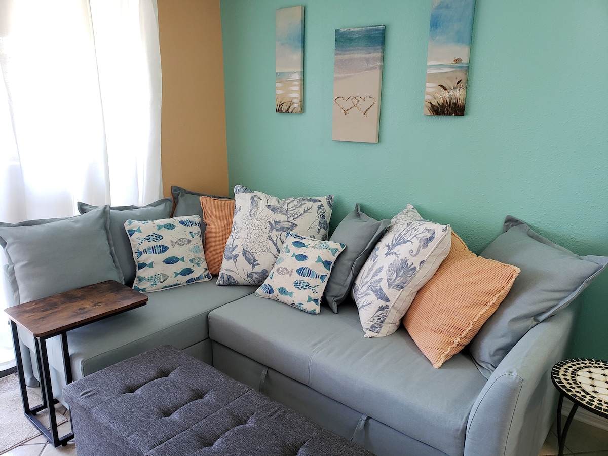 A cozy living area features a sectional sofa adorned with sea-themed cushions. A small wooden table is positioned next to the sofa, while light streams in through sheer curtains, enhancing the calming blue-green wall adorned with coastal artwork.