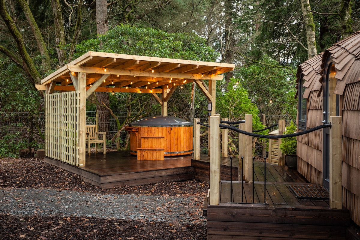 A wooden gazebo with soft lighting surrounds a hot tub, positioned on a deck among lush greenery. A seating area is visible within the gazebo, offering a peaceful retreat. The cabin structure is partially seen on the right, made of natural materials.