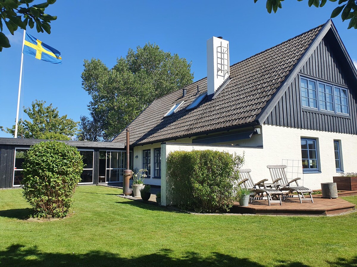 A charming villa features a blend of black and white cladding, framed by lush green grass. Outdoor seating with wooden loungers is positioned on the deck. A large glass sunroom extends the living space, while a Swedish flag proudly waves in the background.