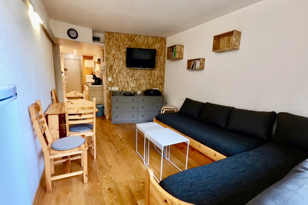 A spacious living area is furnished with a large dark sofa and two wooden tables with matching chairs. A flat-screen TV is mounted on the wall, accompanied by decorative shelving. Natural wood flooring creates a warm ambiance throughout the space.