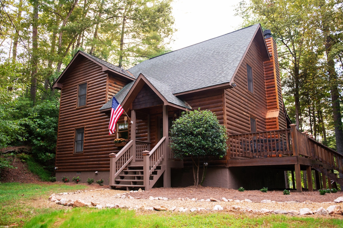 A spacious log cabin is set amidst a forested landscape, featuring a wooden deck and a welcoming entrance. An American flag hangs from the front porch, while shrubs are neatly placed around the exterior. The natural surroundings provide a serene atmosphere.